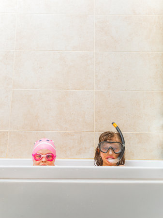 Two cheerful children wearing swim goggles and snorkels peek out from a bathtub. The playful moment takes place against a tiled background, showcasing creativity and delight. Ideal for concepts of fun and innocence.の写真素材