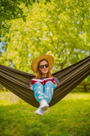 A serene scene of a woman wearing a sunhat and sunglasses, relaxing in a hammock outdoors under the warm sunlight, reading a book surrounded by vibrant green natural scenery.の写真素材