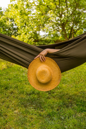 A serene moment of relaxation in a hammock, showcasing a hand holding a straw hat, surrounded by vibrant green nature on a sunny day.の写真素材