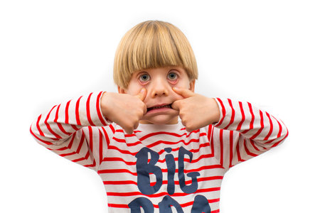 Amusing young child creates a humorous face by pulling on their mouth and eyes while wearing a colorful striped shirt.の写真素材