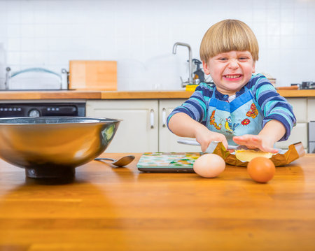 A young child wearing an apron struggles humorously while attempting to bake in a kitchen. The joyful expression and kitchen environment add charm to the playful baking scene.の写真素材