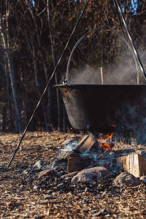 Suspended pot over flames outdoors, set against a wooded background with smoking firewood.の写真素材