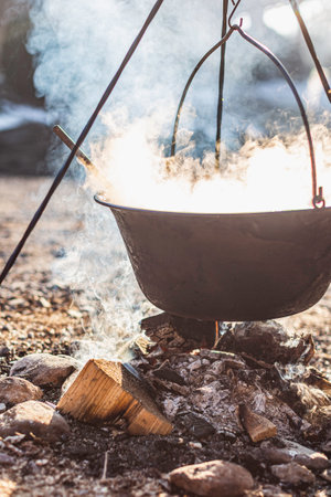 Hanging pot cooking over a smoky fire with wood and ash under open skies.