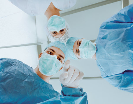 A team of surgeons wearing surgical masks and attire, viewed from the patient's perspective in an operating room. The bright overhead lighting and professional expressions emphasize a moment of medical care and teamwork, symbolizing professionalism, healthcare, and trust in a clinical environment.の写真素材
