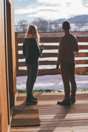 A man and woman stand on a wooden deck, holding wine glasses and looking out at a snowy landscape...