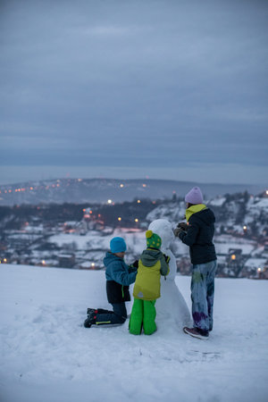 Three children work together to build a snowman on a snowy hill overlooking a town at twilight