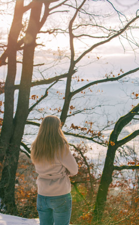 A woman stands with her back to the camera, arms crossed, looking out at a serene winter forest...