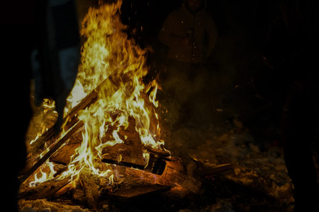 People gather around a large bonfire at night, with flames illuminating the dark surroundings and...