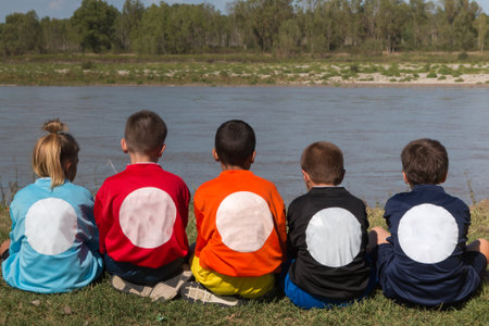 Children in Line Back Turned with Shirts with Different Colors Seated on the Edge of River.