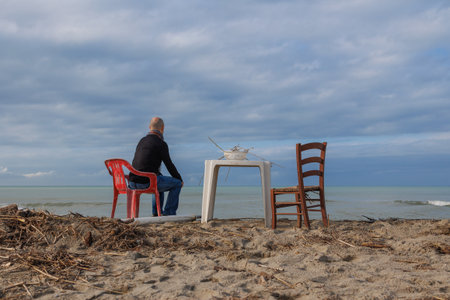 Man Sitting in a Chair Facing the Sea and a Small Table with a Colander resting on it with Wood Remains.の写真素材