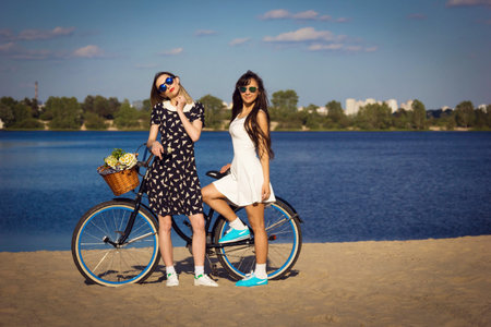 Two beautiful young women on the beach with cruiser bicycle and flowers in basketの写真素材