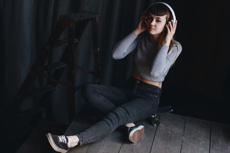 Young girl sitting on skateboard with headphones in loft appartment
