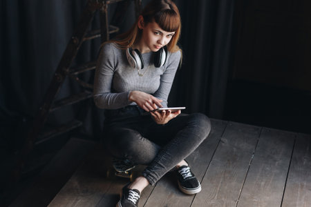 Young girl sitting on skateboard with headphones and tablet in loft appartmentの写真素材