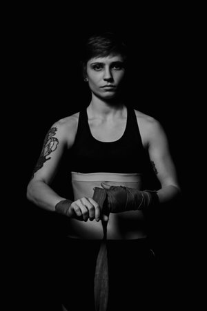 Black and white shot of a young woman wrapping hands with boxing wraps in dark roomの写真素材