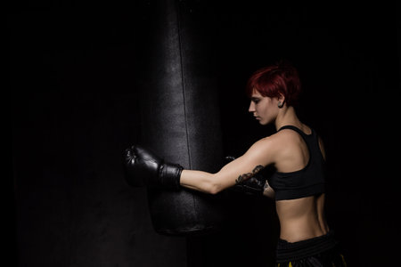 Athlete boxer woman punching a punching bag with dramatic lighting in a dark studioの写真素材