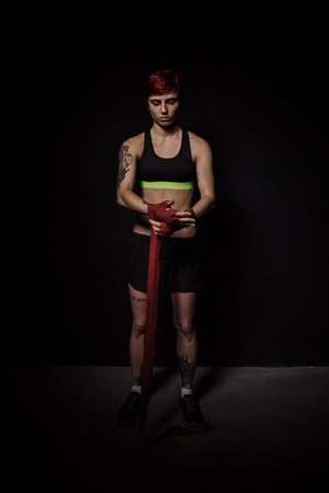 Full length portrait of a young woman wrapping hands with red boxing wraps in dark roomの写真素材