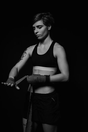 Black and white shot of a young woman wrapping hands with boxing wraps in dark roomの写真素材