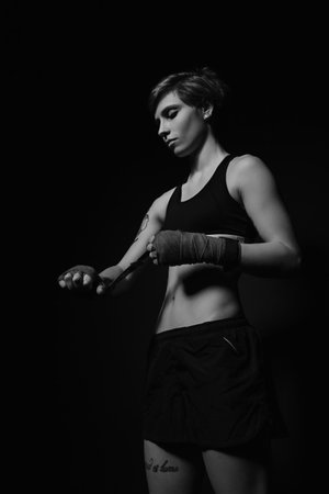 Black and white shot of a young woman wrapping hands with boxing wraps in dark roomの写真素材