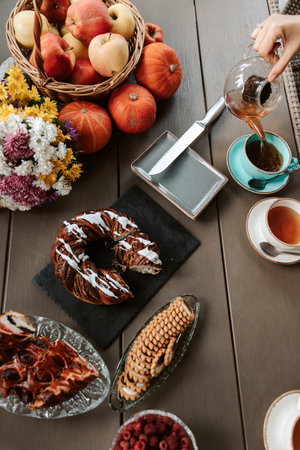 Woman hand pours hot tea from glass teapot into mug. Top view of an autumn table composition with flowers, berries, apples, cookies, pumpkins and pieの写真素材