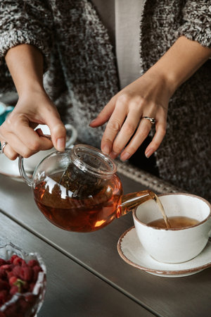 Woman in knitted sweater pours hot tea from glass teapot into mug. Cozy morning breakfast.