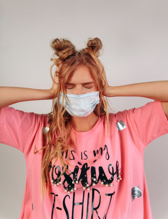 Pretty smiling young european girl dont want to hear holds hands on her ears, keeps eyes closed. Wavy surf hair. Girl with funny hair style. Girl wearing mask isolated on a beige backgroundの写真素材