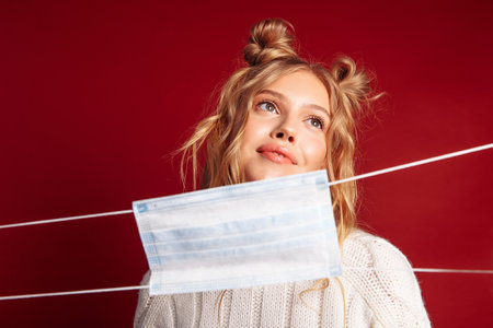 Pretty smiling young european girl ignores wearing mask.  Wavy surf hair. Girl with funny hair style. Girl wearing white sweater, isolated on burgundy background.の写真素材