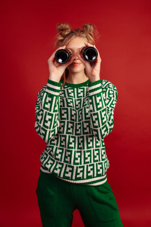 Pretty young smiling girl holds two bottles and simulate that she is looking through binoculars. Wears pullover with green patterns and green pants.  Isolated on a red background. Funny girl.の写真素材