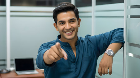 Cheerful young man smiling and pointing at camera in modern office environment, showing confidence, friendliness, and positive energy.の素材