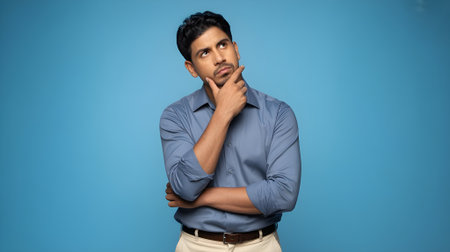 Portrait of a thoughtful young man looking up, representing curiosity, decision-making, and problem-solving against a plain blue background.の素材