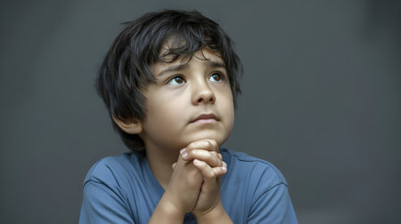 Thoughtful young boy looking up with hands clasped, expressing hope and innocence against a plain background in natural lighting.の素材