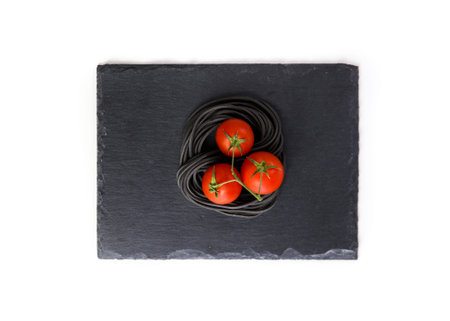 Uncooked black pasta and tomato on a black plateの写真素材