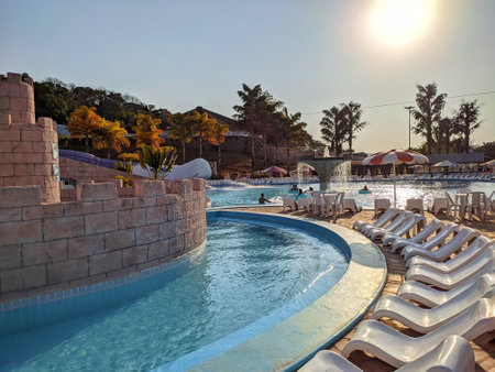 white sun loungers lined up in front of a pool surrounded by chairs and umbrellas with colorful palm trees on a sunny day and blue sky with people in the background at another poolのeditorial素材