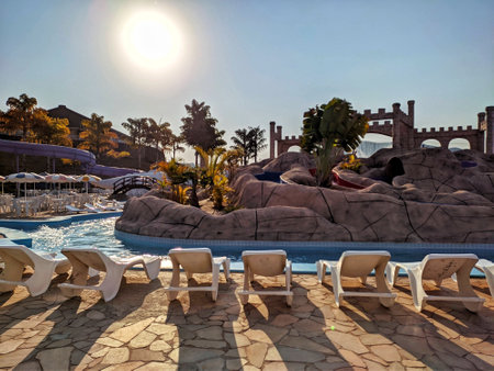 white sun loungers lined up in front of a swimming pool surrounded by chairs and umbrellas with colorful palm trees on a sunny day with blue skiesのeditorial素材
