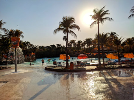 Water park with swimming pool surrounded by palm trees full of people and several beach chairs and umbrellas on a sunny day with blue skyのeditorial素材