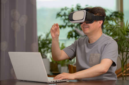 Asian man wearing VR headset and using laptop while sitting at his deskの写真素材