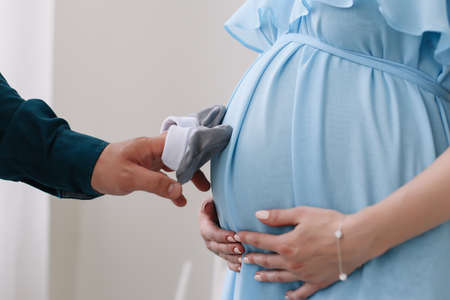 Conceptual photo of couples hands holding baby shoes in anticipation of the child. Pregnant woman in dress on a white background. Pregnancy, maternity, preparation and expectation concept.  Beautiful tender mood photo of pregnancy.の写真素材