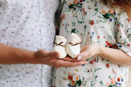 Conceptual photo of couples hands holding baby shoes in anticipation of the child. Pregnant woman in dress on a white background. Pregnancy, maternity, preparation and expectation concept.  Beautiful tender mood photo of pregnancy.の写真素材