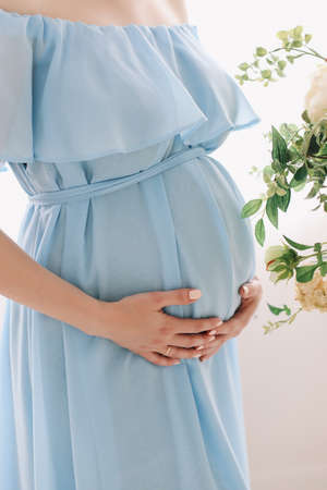 Pregnant woman in dress holds hands on belly on a white background. Pregnancy, maternity, preparation and expectation concept. Close-up, indoors. Beautiful tender mood photo of pregnancy.の写真素材