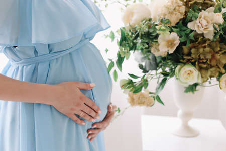 Pregnant woman in dress holds hands on belly on a white background. Pregnancy, maternity, preparation and expectation concept. Close-up, indoors. Beautiful tender mood photo of pregnancy.の写真素材