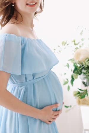 Pregnant woman in dress holds hands on belly on a white background. Pregnancy, maternity, preparation and expectation concept. Close-up, indoors. Beautiful tender mood photo of pregnancy.の写真素材
