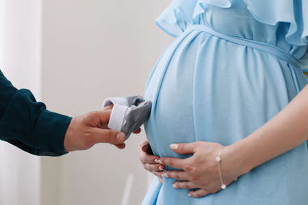 Conceptual photo of couples hands holding baby shoes in anticipation of the child. Pregnant woman in dress on a white background. Pregnancy, maternity, preparation and expectation concept.  Beautiful tender mood photo of pregnancy.の写真素材