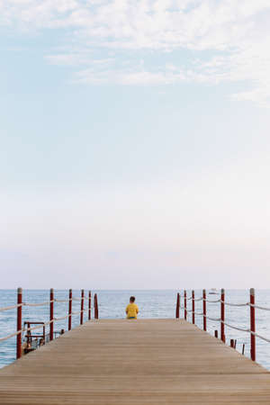 A boy sitting on the pier and looking at the sea horizon and blue sky. Vacation concept. Summer time sea vacation backgroundの写真素材