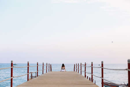 Girl sitting on the pier and looking at the sea horizon and blue sky. Vacation concept. Summer time sea vacation backgroundの写真素材