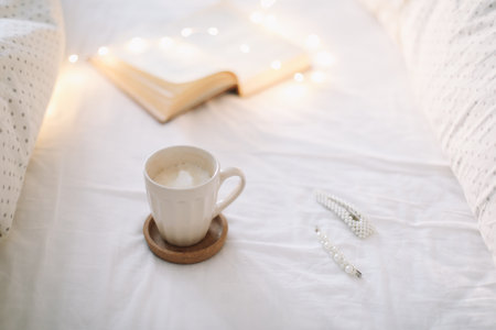 Coffee cup with milk foam and a book on a white bedsheet.の写真素材