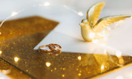 Golden wedding rings on golden background with toy bird and decorations. Shiny golden jewelry. Wedding details. Proposal of marriage, valentine gift. copy space.の写真素材