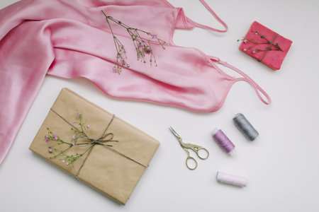 Composition with pink silk fabric, gift in craft paper and threads and scissors on white background. Flat lay, top view. Hobby, leisure concept.の写真素材