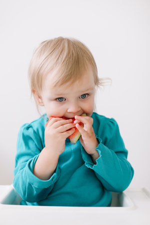 portrait of a funny little baby girl sitting in a high chair and eating watermelon