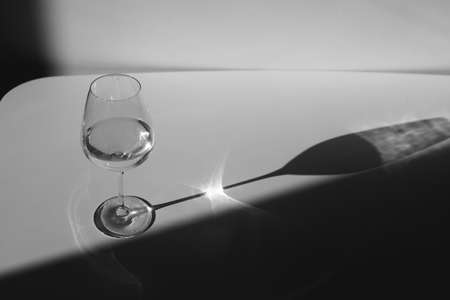 A glass of water on a white background with a shadow. Chiaroscuro from the window. Black and white photo. Close-up, copy spaceの写真素材