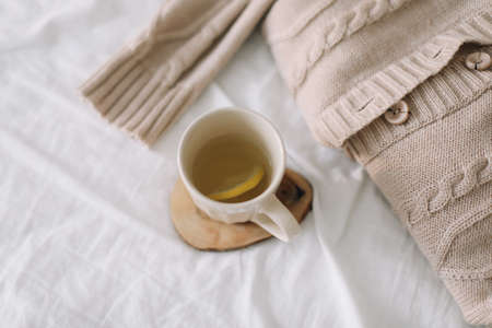 Knitted beige sweater and cup of tea on messy bed background. Winter and spring fashion concept. Top...