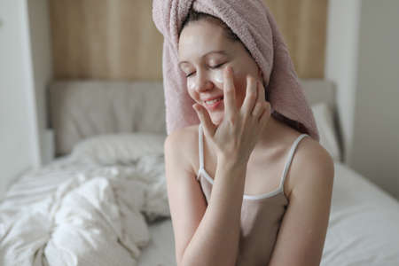 smiling young woman with a towel on head apply cream on face skinの写真素材